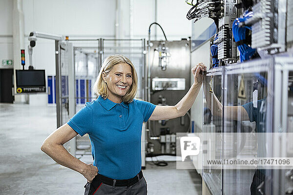 Lächelnde Berufstätige mit der Hand auf der Hüfte  die an Maschinen in der Fabrik steht