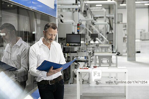 Mature businessman reading file while leaning on glass in factory