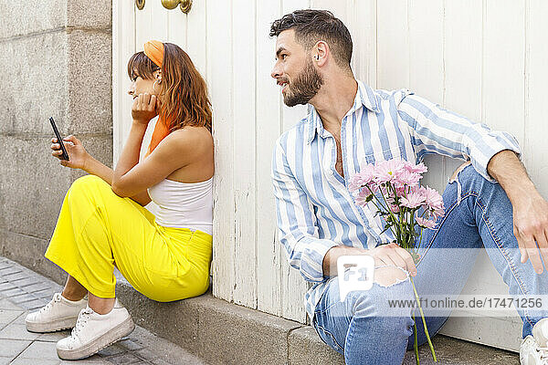 Young man looking at woman using mobile phone while sitting at doorway