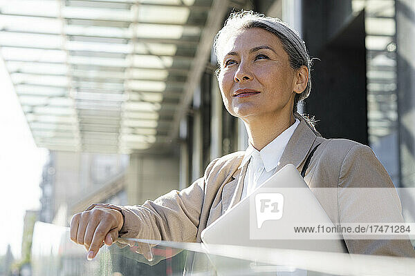 Thoughtful businesswoman with laptop standing by glass railing