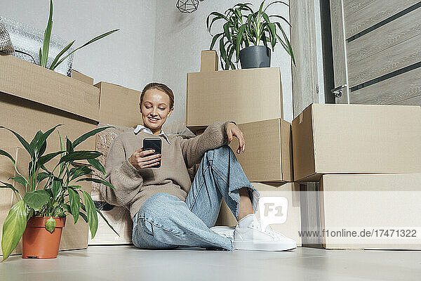 Smiling young woman using mobile phone in front of cardboard boxes in new home