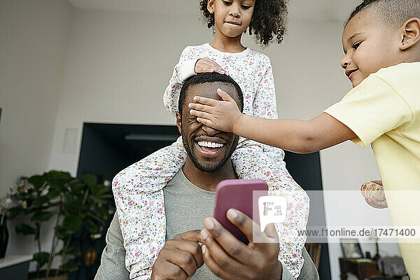 Playful boy and girl having fun with father holding smart phone at home