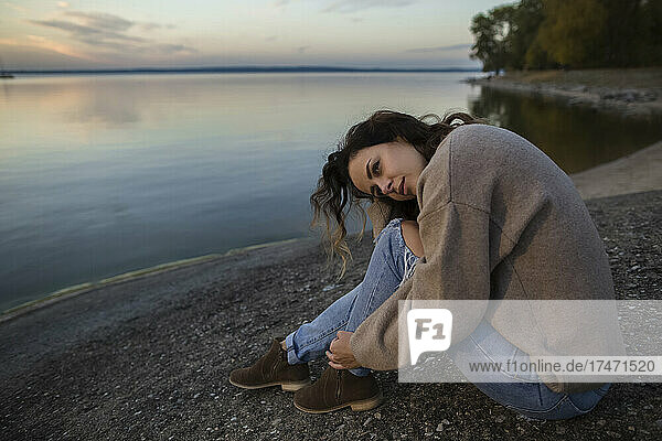 Thoughtful woman sitting on rock by lake at sunset