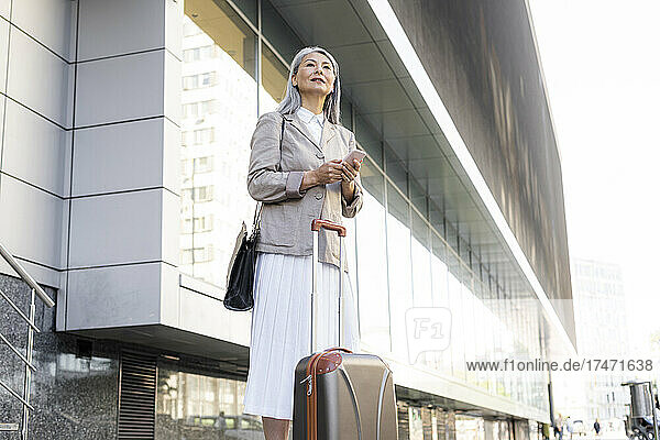 Geschäftsfrau mit Koffer hält Smartphone in der Hand  während sie vor dem Bürogebäude steht