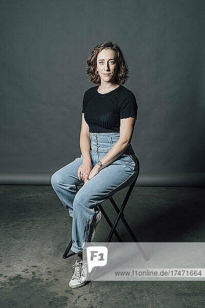 Confident woman sitting on chair in front of gray backdrop