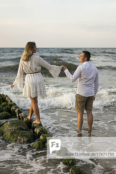 Girlfriend holding hand of boyfriend while walking on rock in sea