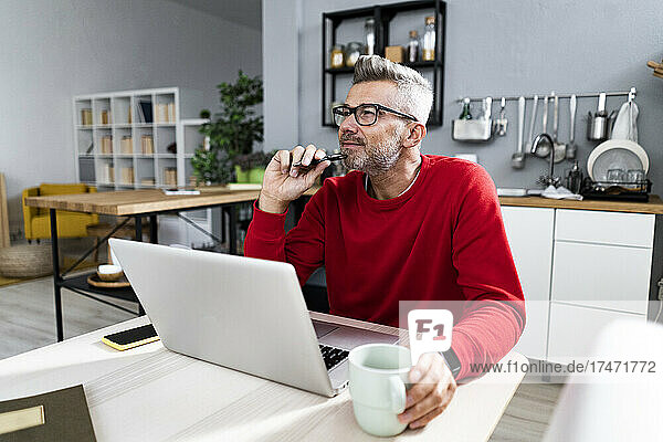 Thoughtful man wearing eyeglasses sitting with laptop at table