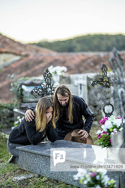 Mann tröstet Frau am Grabstein auf dem Friedhof