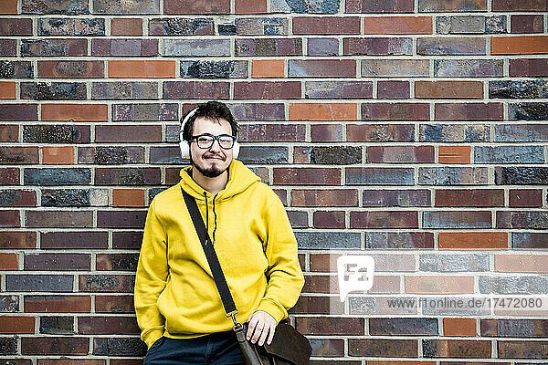 Smiling student with bag listening music in front of brick wall