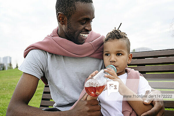 Smiling father looking at boy drinking juice on bench