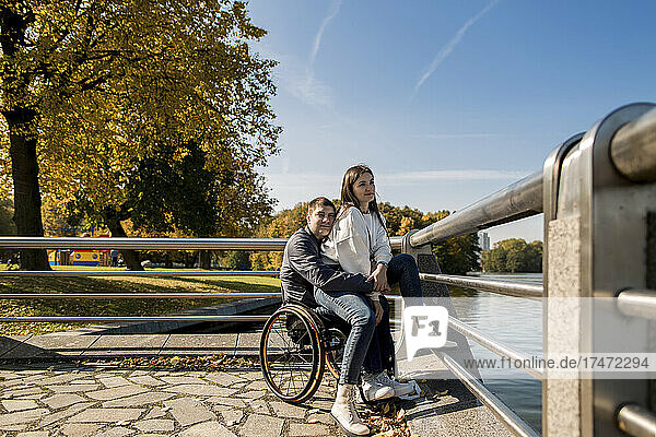 Girlfriend sitting with disabled boyfriend in wheelchair at embankment