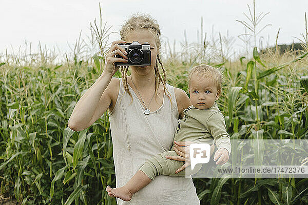 Mother photographing through camera carrying toddler girl in front of crops