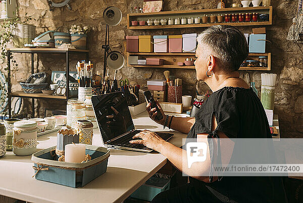 Mature female artist with laptop using mobile phone in workshop