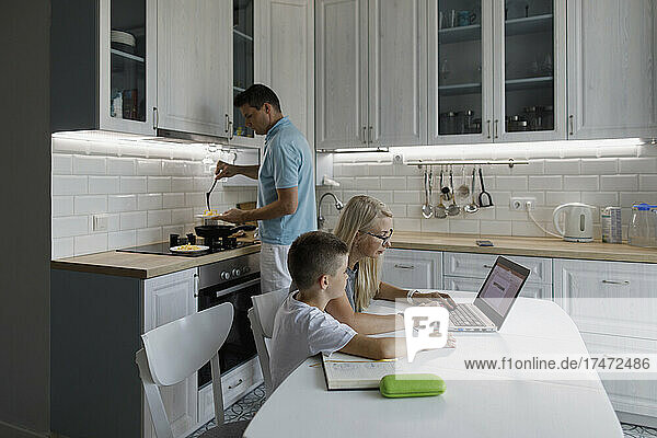 Mother helping son with homework on laptop at table by man cooking in kitchen