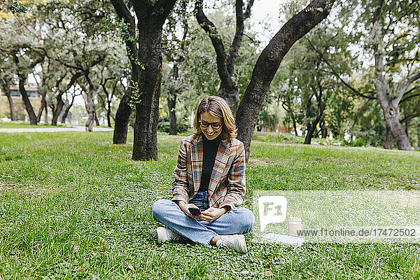 Lächelnde Geschäftsfrau  die Textnachrichten per Smartphone auf Gras im Park sendet