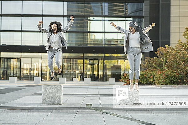 Cheerful female colleagues jumping on footpath in front of building