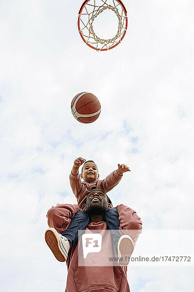 Son throwing basketball towards hoop while sitting on father's shoulders