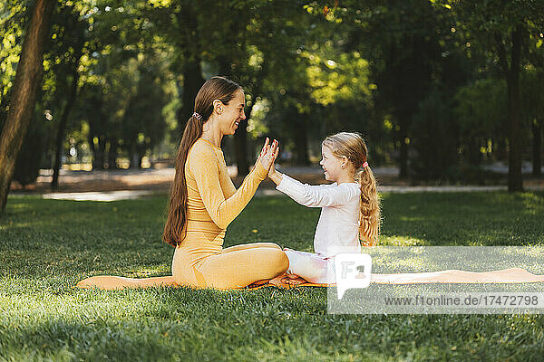 Smiling daughter touching mother's hands on exercise mat in park