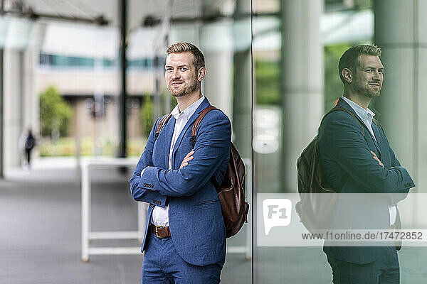 Smiling businessman standing with arms crossed by glass wall