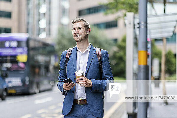 Smiling businessman holding disposable cup and mobile phone in city