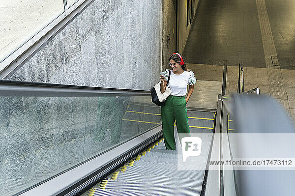 Young female professional standing on escalator listening music using smart phone