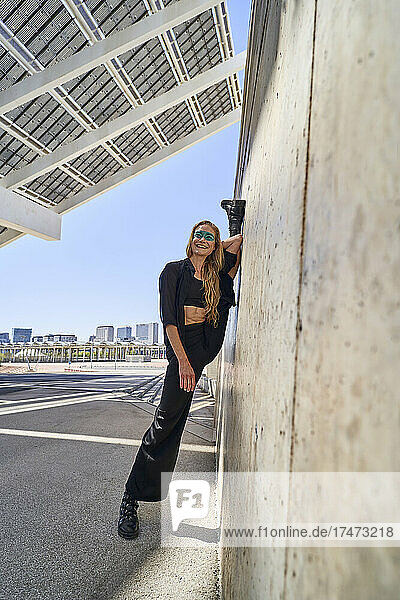 Smiling woman with leg up by wall on sunny day