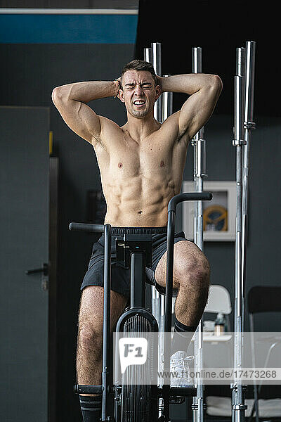 Tired male athlete with hands behind head sitting on cross trainer in gym