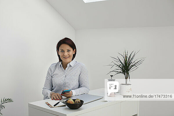 Smiling freelancer with smart phone in front of while wall at home office