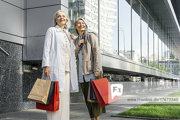 Smiling women looking away while holding shopping bags outside mall