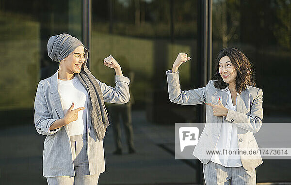 Smiling female professionals pointing while flexing muscles in front of glass wall