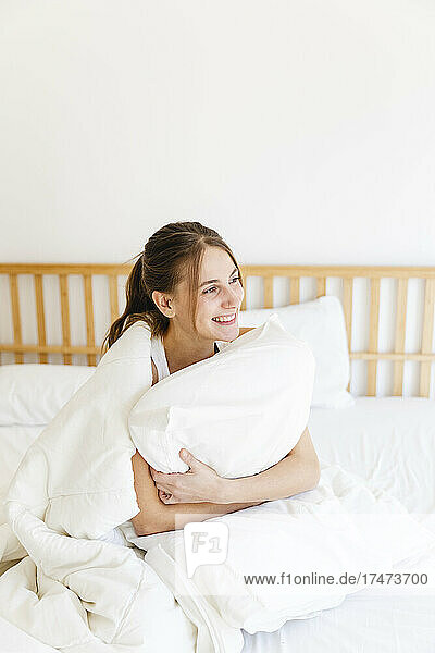 Lächelnde junge Frau umarmt Kissen  während sie im Schlafzimmer auf dem Bett sitzt