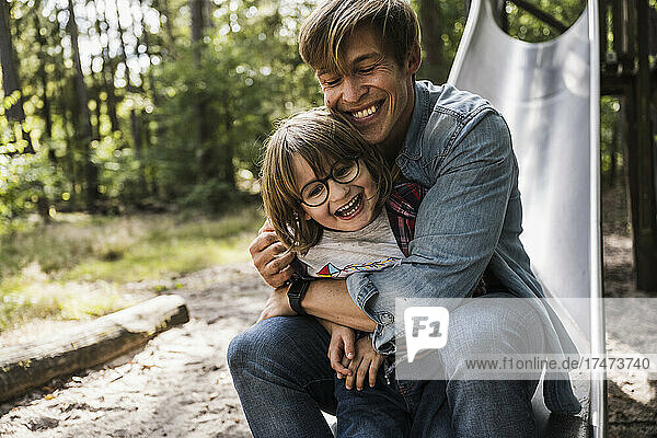 Smiling father with eyes closed embracing son on slide in forest