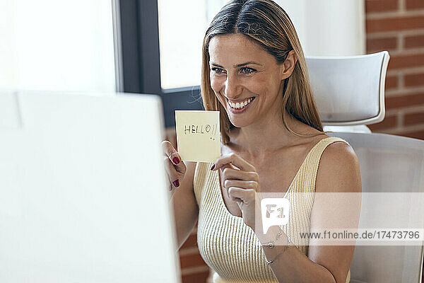 Smiling female freelancer showing adhesive note during video conference through computer at home