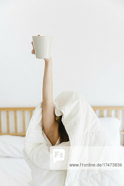 Woman with hand raised holding coffee cup while wrapped in white blanket on bed