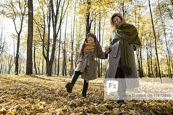Smiling woman holding hand of daughter kicking autumn leaves in park