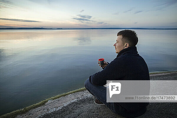 Man drinking tea sitting on rock by lake at sunset