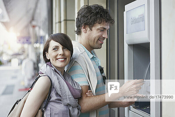 Smiling man withdrawing money from ATM standing by girlfriend