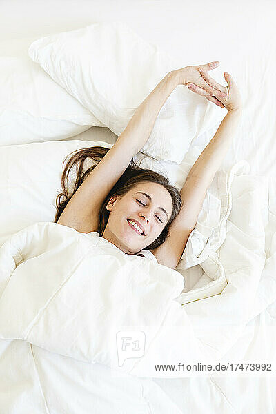 Smiling young woman stretching while lying with while blanket in bedroom