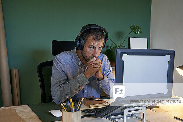 Thoughtful businessman wearing headphones looking at laptop in home office