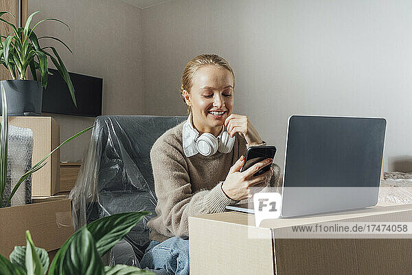 Smiling woman with laptop on carton using mobile phone in relocated home
