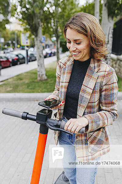 Smiling freelancer unlocking push scooter through mobile phone outside park