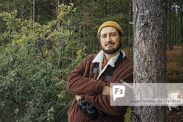 Man with binoculars leaning on tree in forest