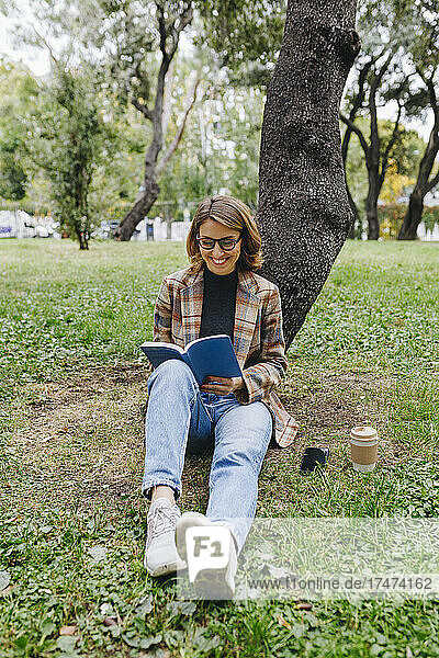 Smiling freelancer reading book in front of tree in park