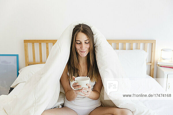 Junge Frau mit braunem Haar hält eine Tasse Kaffee auf dem Bett