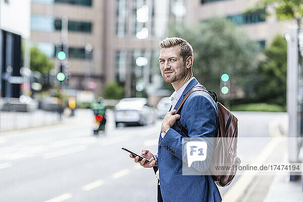 Young businessman with backpack holding smart phone in city