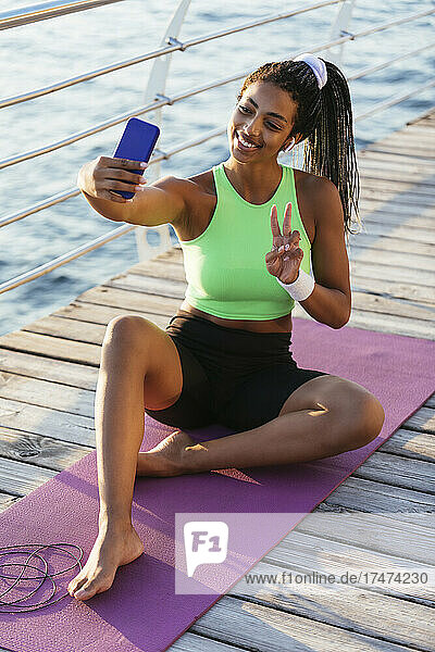 Smiling young sportswoman gesturing peace sign while taking selfie through smart phone
