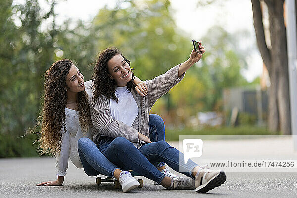 Smiling woman taking selfie with female friend through smart phone while sitting on skateboard