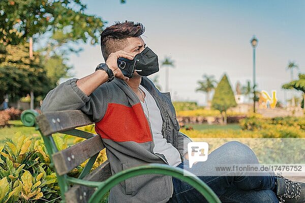 Mann sitzt auf einer Bank und telefoniert  junger Mann mit Maske telefoniert mit dem Handy  Mann mit Maske telefoniert in einem Park  Nicaragua  Mittelamerika