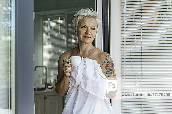 Woman with tattoo wrapped in sheet drinking coffee