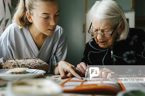 Mitarbeiterin des Gesundheitswesens löst Kreuzworträtsel mit einer älteren Frau  während sie zu Hause in der Küche sitzt